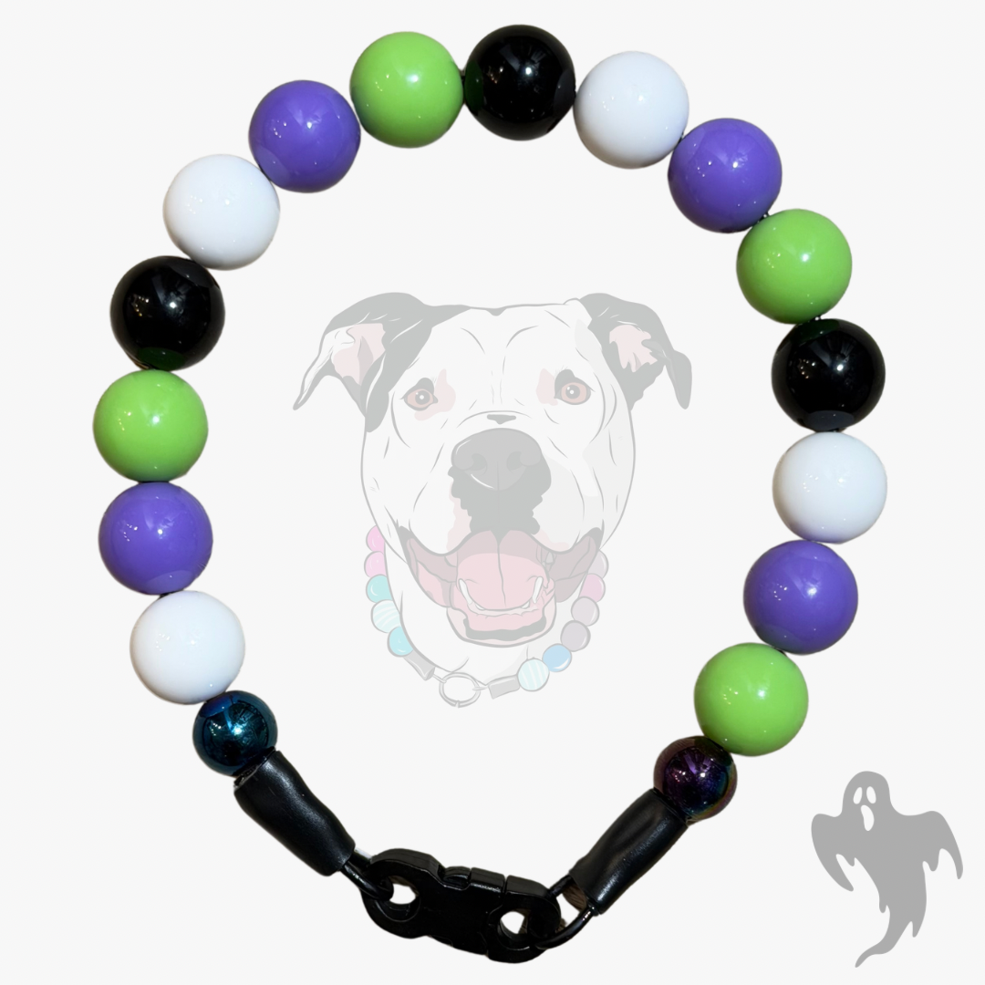 A beaded dog collar with alternating glossy beads in purple, green, black, white, and one iridescent blue bead, separated by black spacer beads and a black clasp. A faint dog face graphic is visible in the background.
