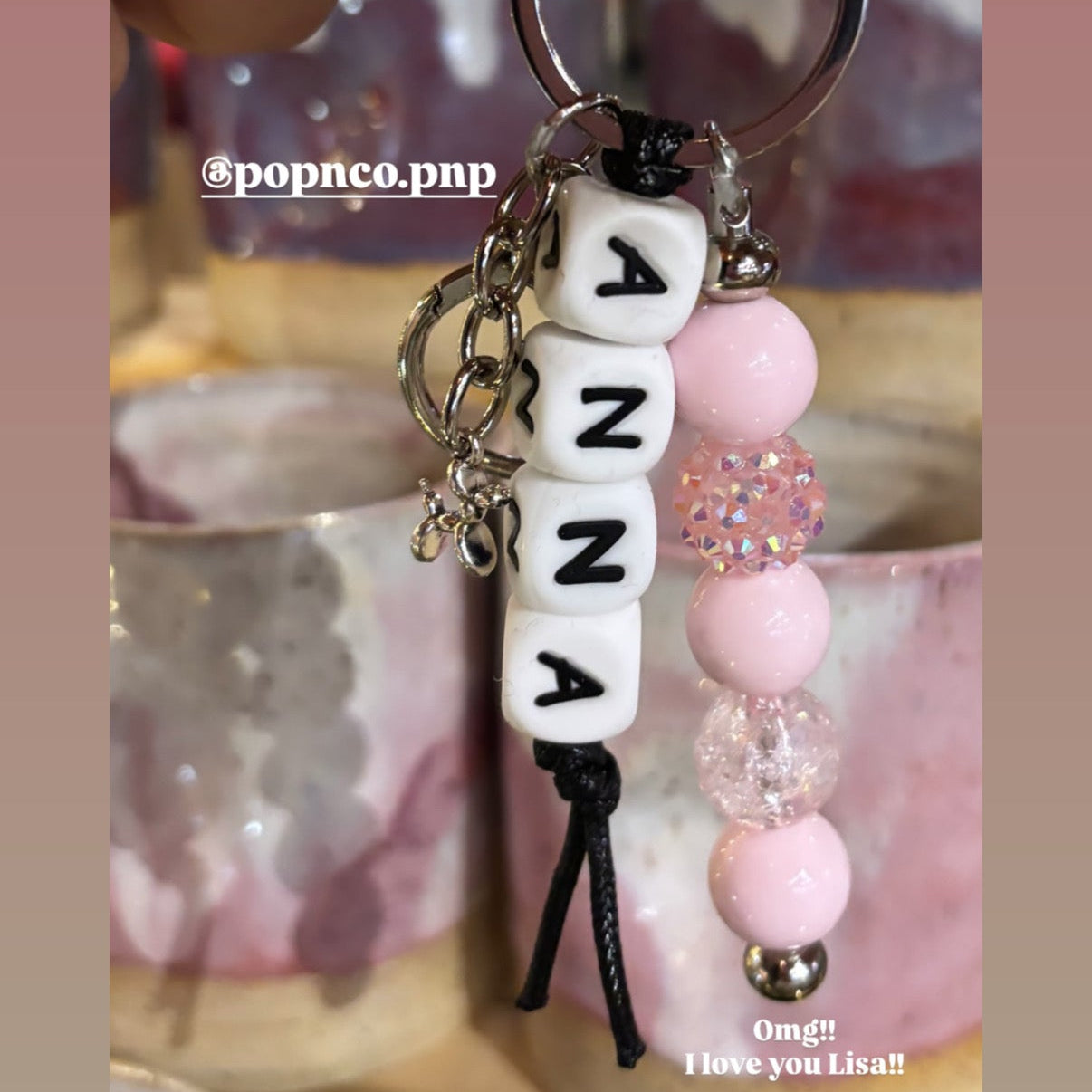 Keychain with 'ANNA' beads held over a blurred background POPNCO