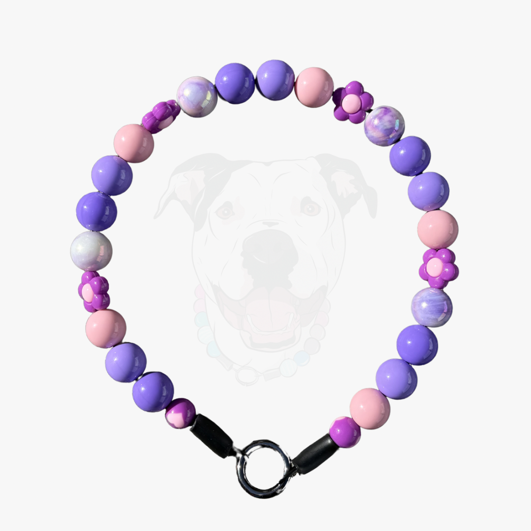 A beaded dog collar featuring purple and pink round beads, marbled beads, small flower-shaped beads, and a black metal ring clasp, shown against a light gray background with a faint dog graphic.