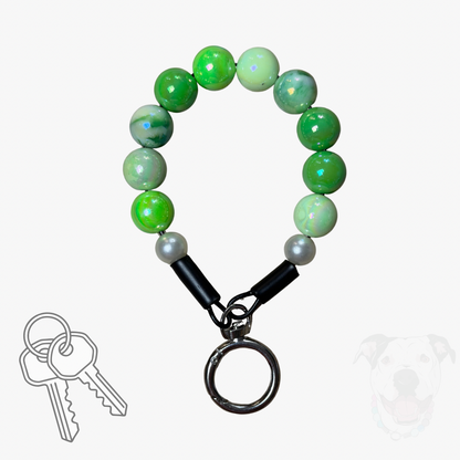 A beaded keychain with green marbled and light green beads, black connector caps, and a silver metal key ring, shown on a white background with small decorative graphics of keys and a dog silhouette.