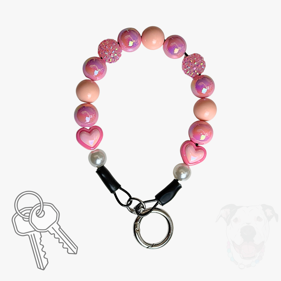 Pink beaded dog leash keychain with heart and glitter beads, a silver split ring, and a black snap clasp with two keys, shown on a white background with a faint dog silhouette and key icon.