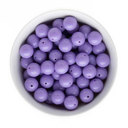 A top-down view of light purple lavender round beads with small holes, arranged inside a white ceramic bowl on a white background.
