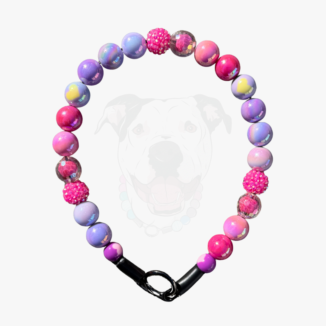 A beaded dog collar with pink, purple, and blue marbled beads, textured pink pom-pom beads, and a black clasp, shown against a light gray background with a faint dog silhouette.