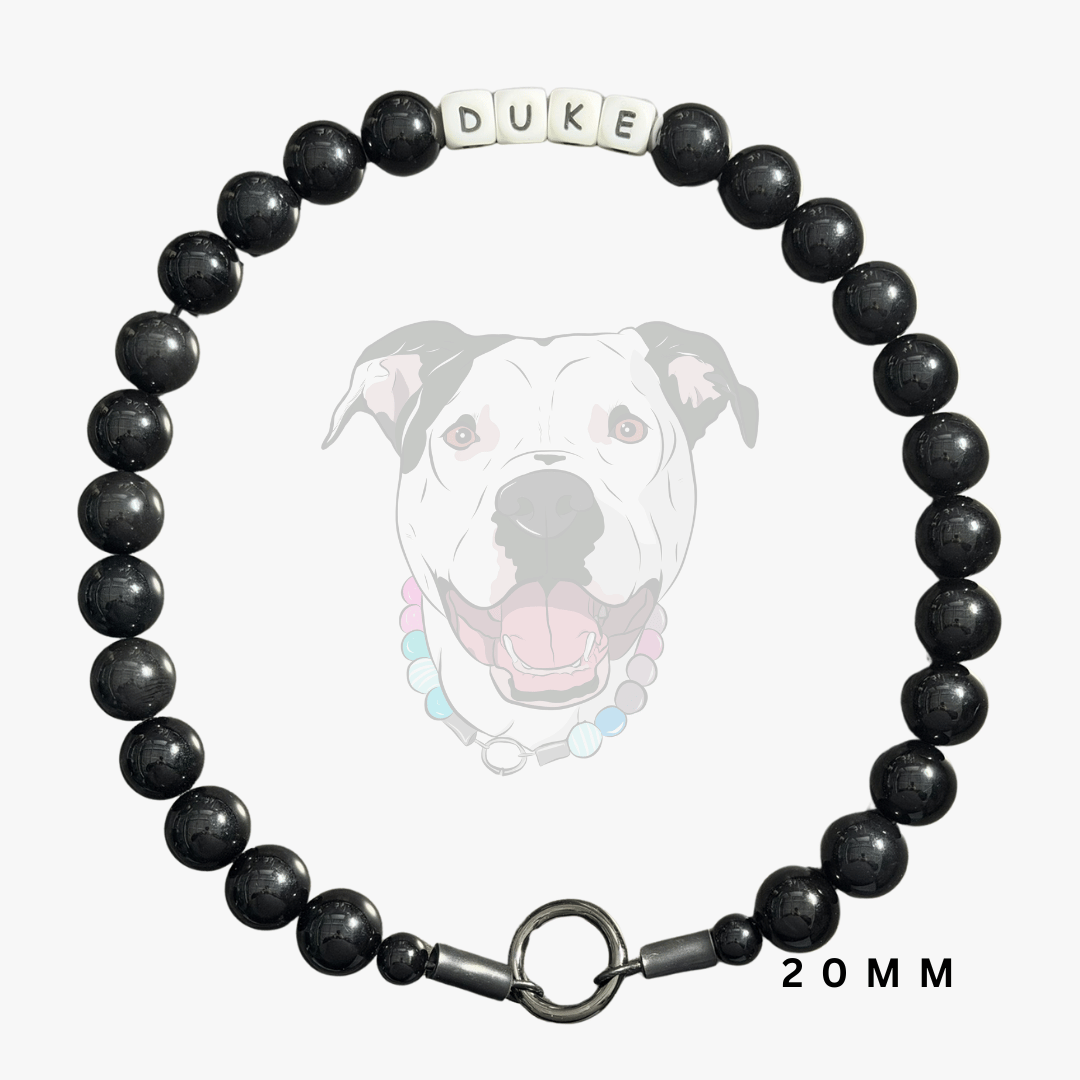 A black beaded dog collar with a silver clasp and a white letter bead spelling 'DUKE', shown on a light gray background with a faded dog illustration and '20MM' text.