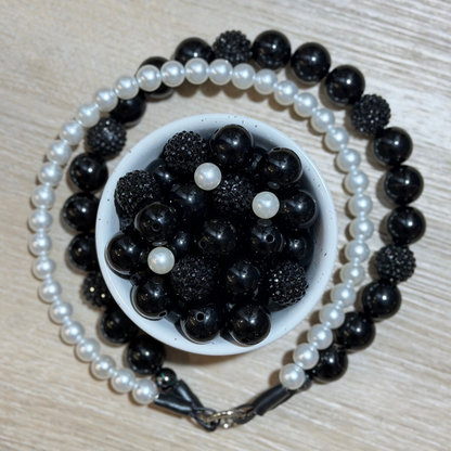 Black and white beaded pet collar arranged in a circle around a small white bowl filled with black beads and a few white pearl-like beads, on a light wood surface.