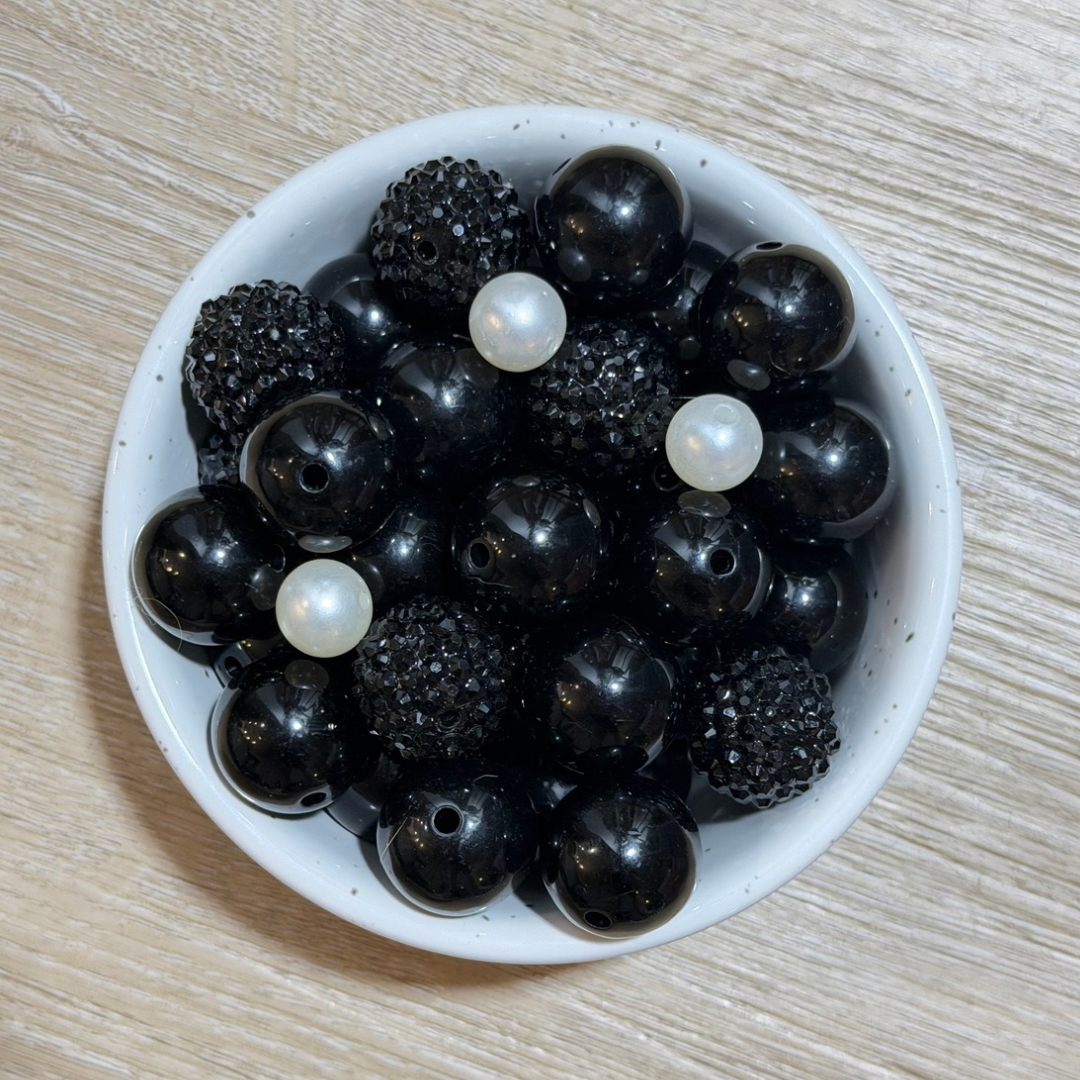A top-down view of a white speckled bowl filled with black round beads, including some smooth glossy black beads and some covered in small clear rhinestones, with a few white faux-pearl beads on top, set on a light wood surface.