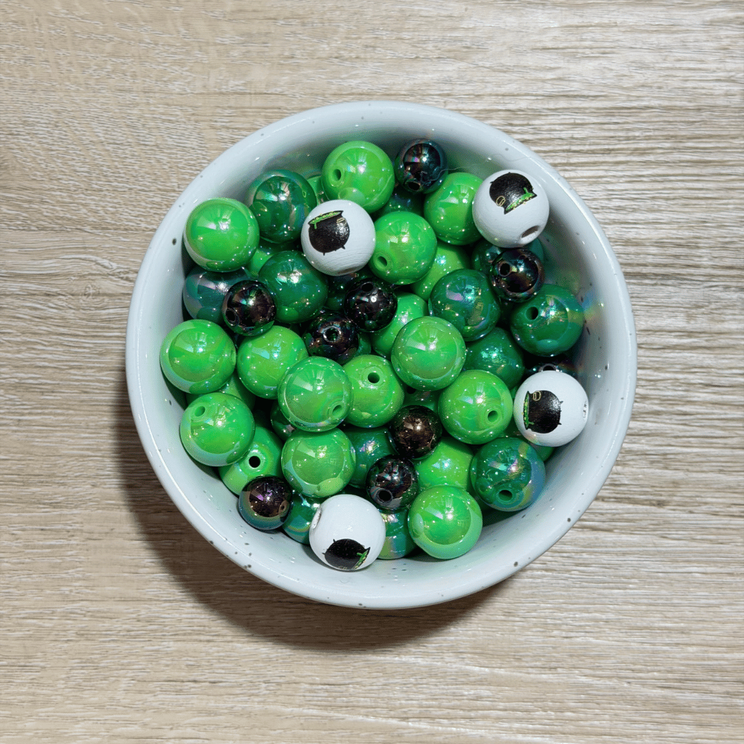 A top-down view of a white ceramic bowl filled with green, black, and white eyeball beads, set on a light wood grain surface.