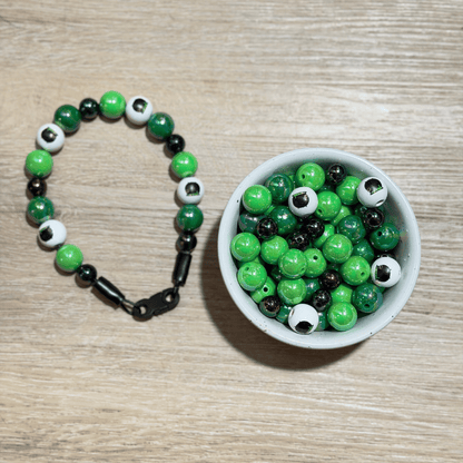 A beaded bracelet made of green, black, and white evil eye beads is displayed next to a small white bowl filled with the same beads on a light wood background.