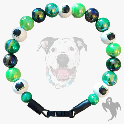 A beaded dog collar featuring green, white, and iridescent blue-green beads, with one white bead printed with a black haunted-house silhouette. The collar is displayed against a light gray background with a faded graphic of a dog wearing the collar.