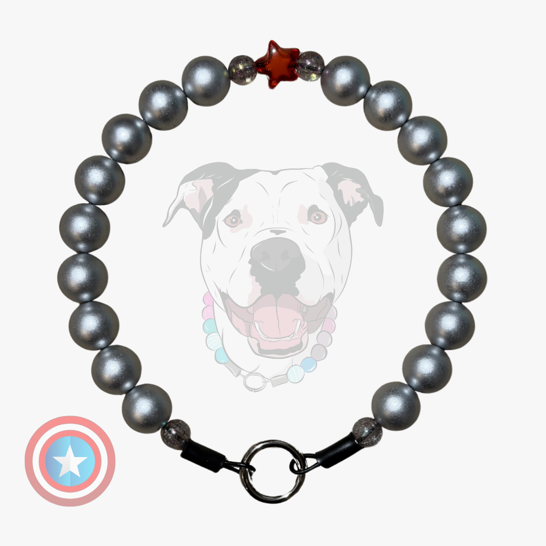 A beaded dog collar featuring large, glossy dark gray pearls, a small red star-shaped bead, gray spacer beads, and a black toggle clasp. A faint graphic of a white pitbull dog is visible in the background, and a Captain America shield logo is in the bottom-left corner.