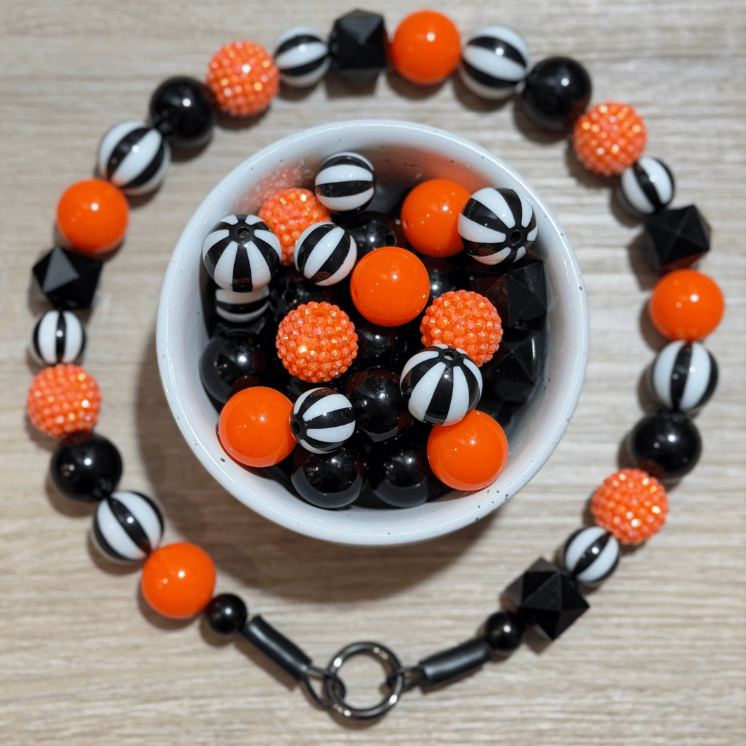 A beaded bracelet arranged in a circle on a light wood surface, with a small white bowl in the center filled with matching beads. The bracelet features bright orange, glossy black, and black-and-white striped round beads, with textured orange and black faceted beads.