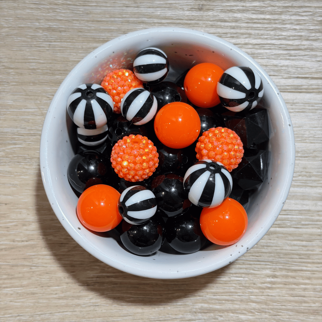 A white ceramic bowl filled with decorative balls in black, white, orange, and textured orange, placed on a light wood surface.