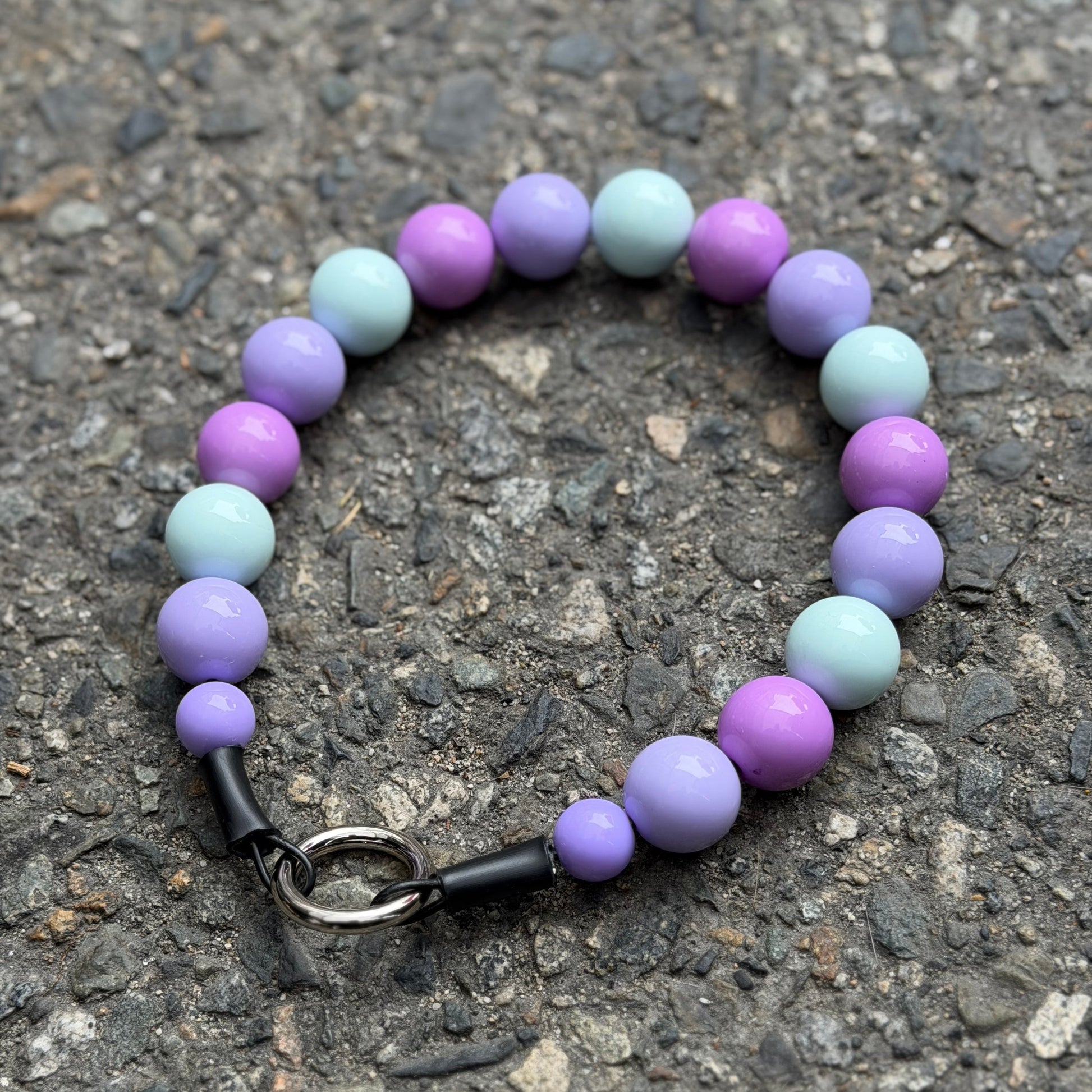 A beaded pet collar with alternating light purple, lavender, and pale mint-green beads, featuring an O ring slip on style and made on Aircraft Grade Stainless Steel Wire, photographed on a textured gray asphalt surface 