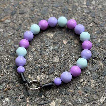 A beaded pet collar with alternating light purple, lavender, and pale mint-green beads, featuring an O ring slip on style and made on Aircraft Grade Stainless Steel Wire, photographed on a textured gray asphalt surface 