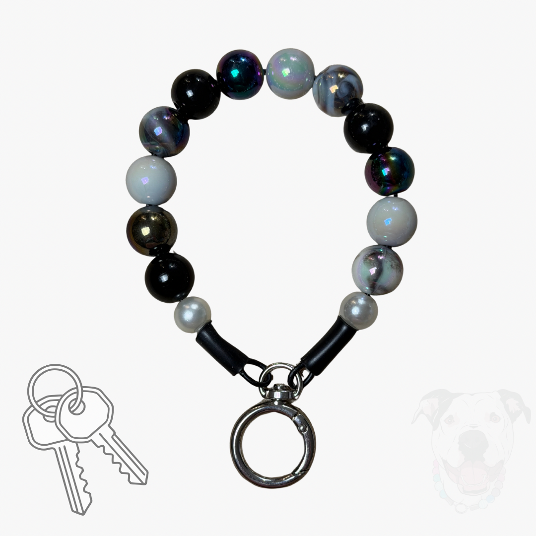 A beaded dog collar with black, white, and iridescent beads, featuring a silver metal key ring clasp. A set of keys is shown in the bottom-left corner, and a faint dog silhouette is visible in the bottom-right corner, all on a light gray background.