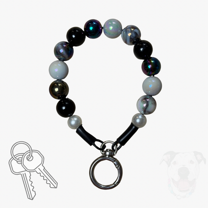 A beaded dog collar with black, white, and iridescent beads, featuring a silver metal key ring clasp. A set of keys is shown in the bottom-left corner, and a faint dog silhouette is visible in the bottom-right corner, all on a light gray background.