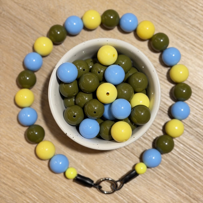 A beaded necklace made of glossy round beads in light blue, olive green, and pale yellow, arranged around a small white bowl filled with loose beads of the same colors, on a light wood surface.