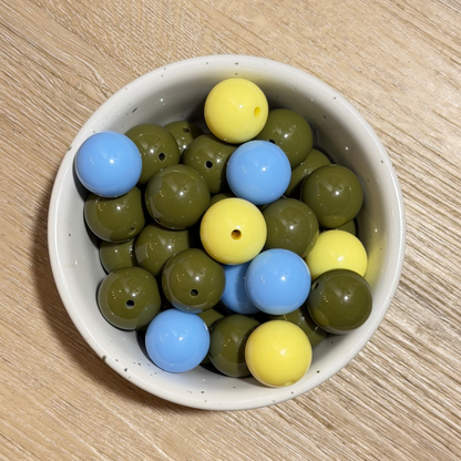 A bowl of round wooden beads in olive green, light blue, and pale yellow, arranged on a light wood tabletop.