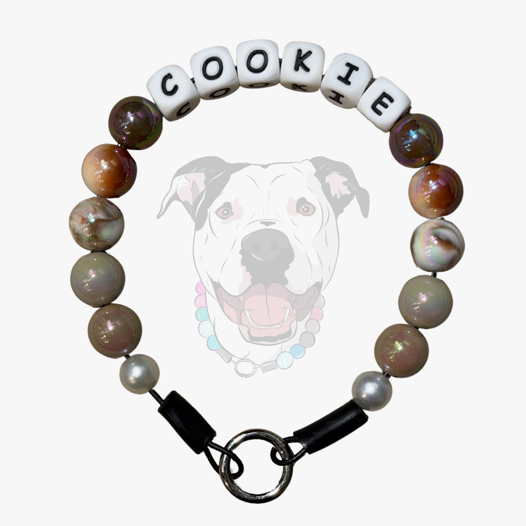 A beaded dog collar featuring white letter beads spelling 'COOKIE' and marbled brown, gray, and white beads, with a black clasp and metal ring, shown against a light gray background with a faint dog graphic.