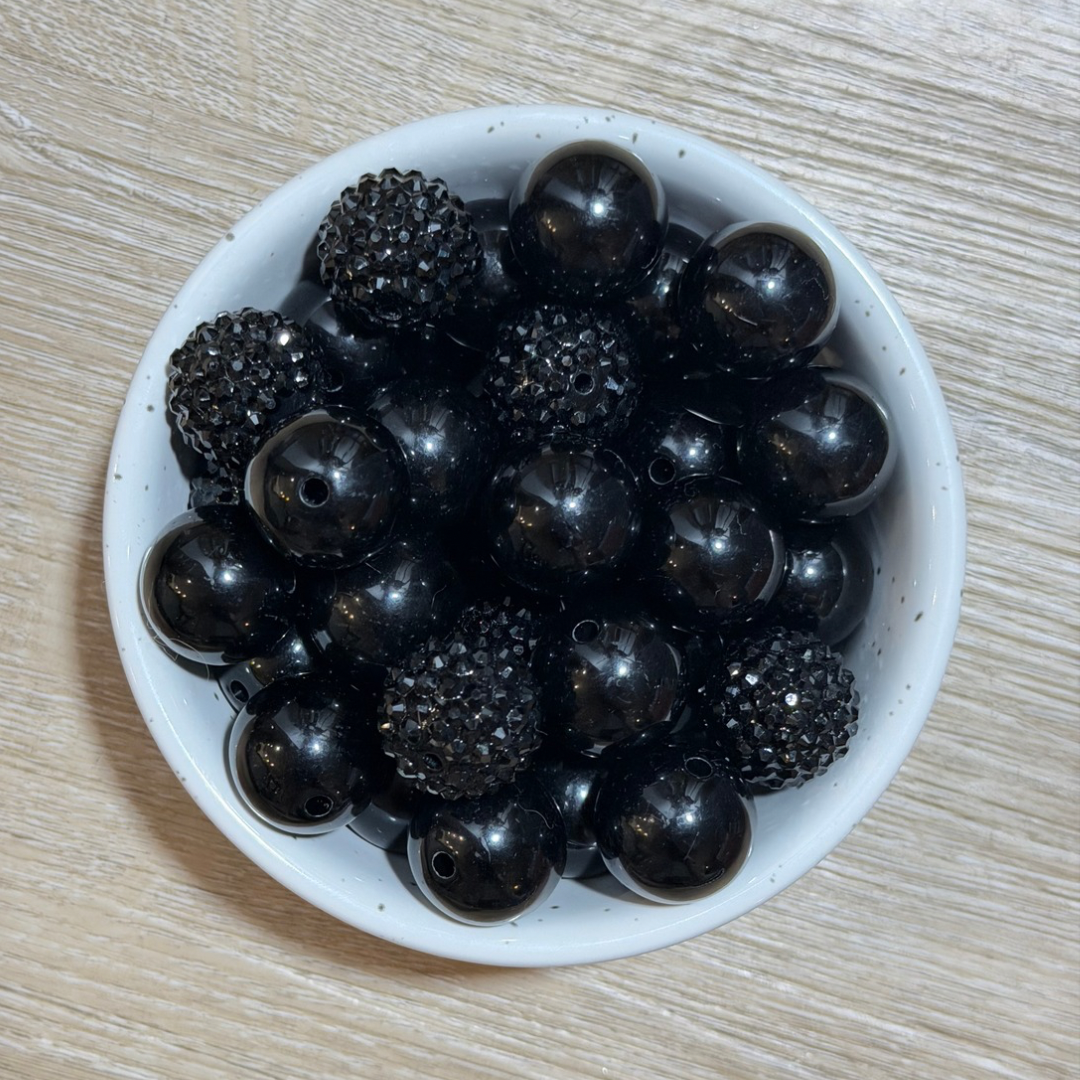 A top-down view of glossy black seed beads, including smooth round beads and textured spiky dome beads, arranged in a small white speckled ceramic bowl on a light wood surface.