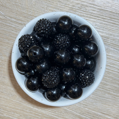 A top-down view of glossy black seed beads, including smooth round beads and textured spiky dome beads, arranged in a small white speckled ceramic bowl on a light wood surface.