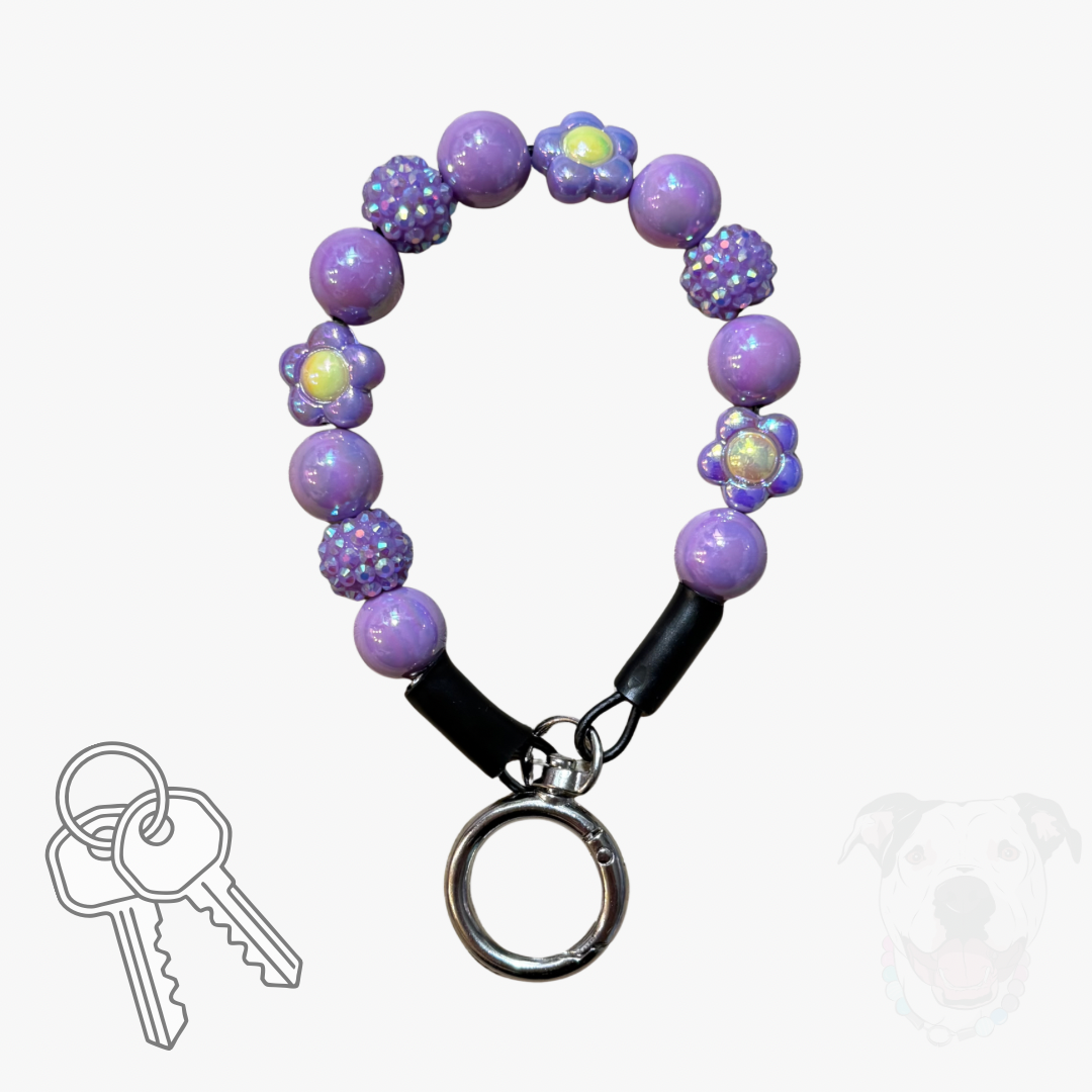 A purple beaded keychain with flower and textured bead details, a black leather strap, and a silver metal key ring, shown on a light gray background with a small key graphic and a faint dog silhouette.