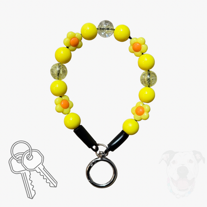Yellow beaded dog collar keychain with flower charms, clear textured beads, black toggle clasp, and attached key ring with keys, shown on a light gray background with a faint dog silhouette and key icon.