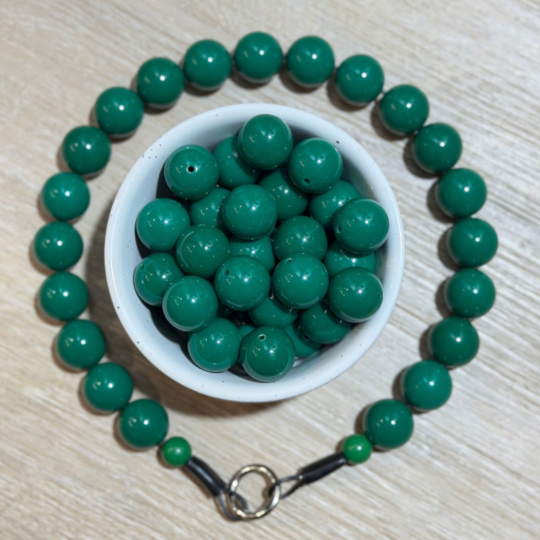 Green round bead necklace arranged in a circle around a small white bowl filled with matching green beads, on a light wood surface.
