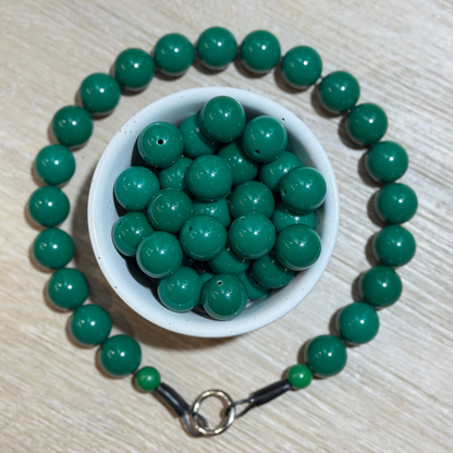 Green round bead necklace arranged in a circle around a small white bowl filled with matching green beads, on a light wood surface.
