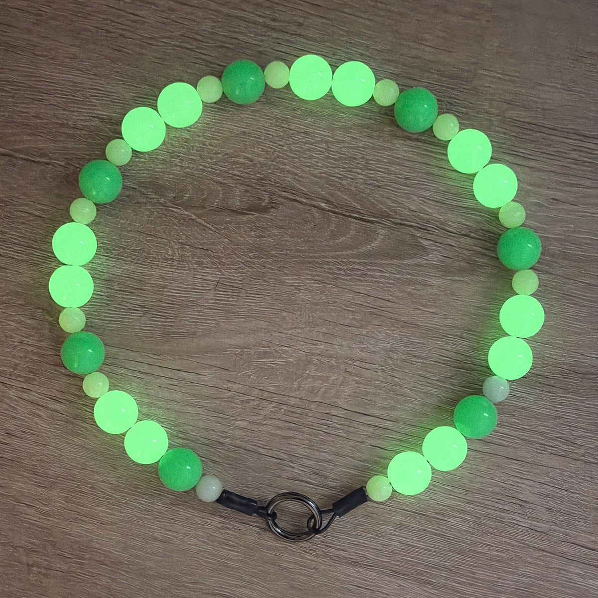 A beaded necklace made of glowing neon green and light green round beads, with a black cord and metal ring clasp, laid flat on a brown wooden surface.