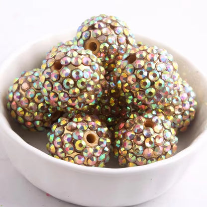 A pile of round rhinestone-covered beads with iridescent, rainbow-like reflections, displayed in a small white bowl against a light background.