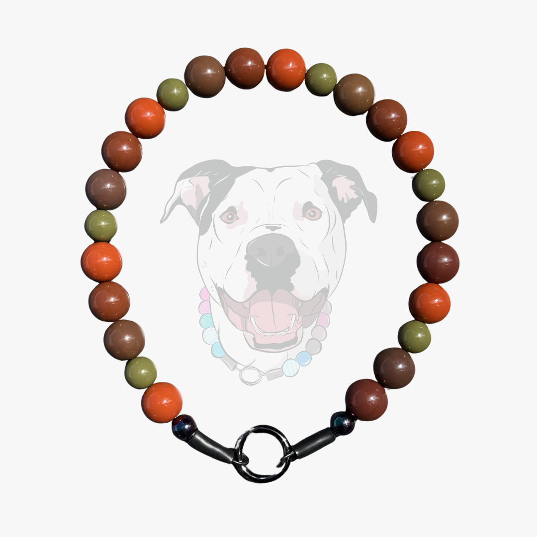 A beaded dog collar necklace featuring alternating brown, orange, and olive green round beads, finished with a black metal ring clasp, shown against a light gray background with a faint graphic of a pitbull dog.