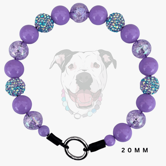 A purple beaded pet collar shown on a light gray background with a faded dog graphic behind it. The collar features smooth purple beads, faceted purple beads filled with glitter, and iridescent blue rhinestone-covered beads, finished with a black clasp and silver O-ring.