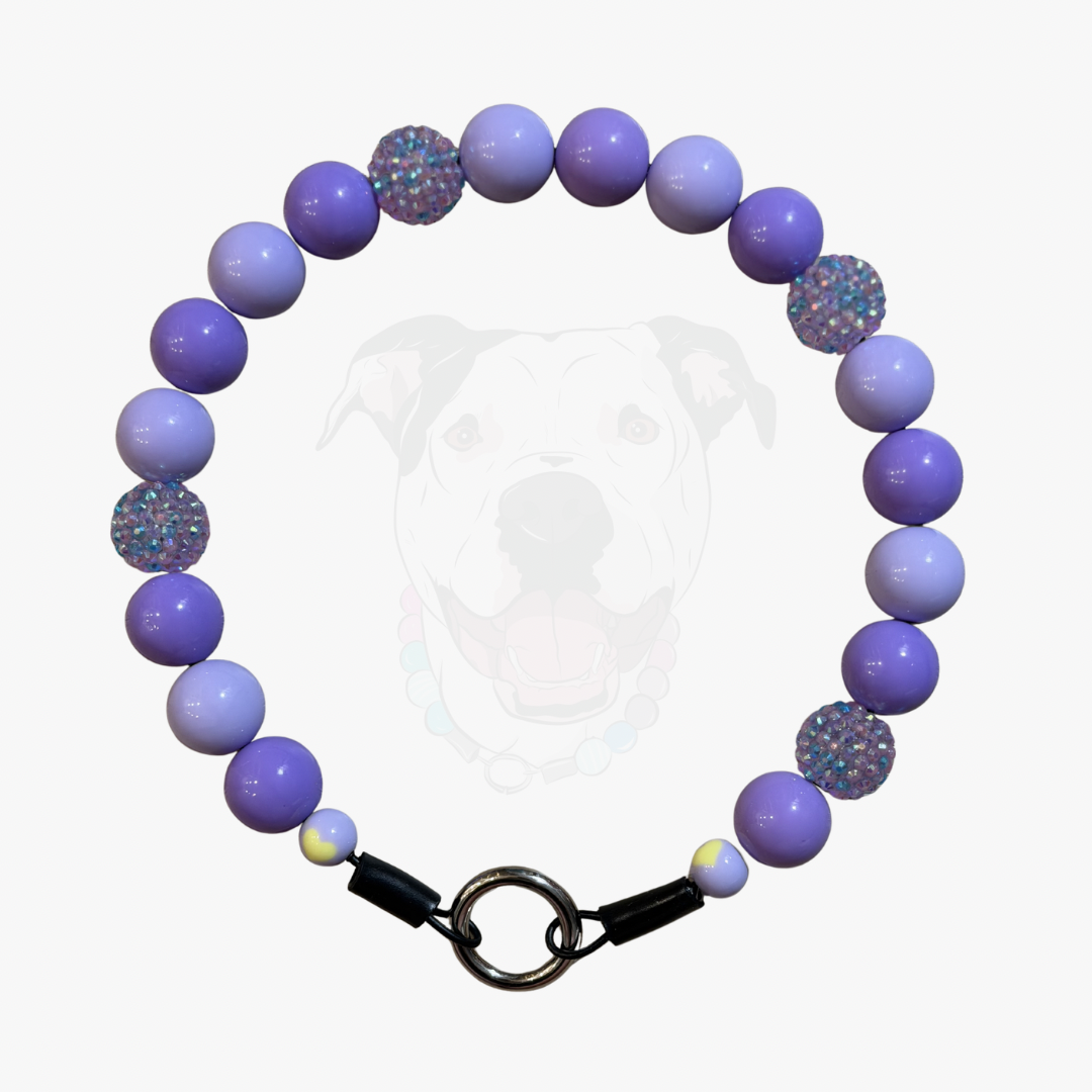 A beaded pet collar featuring light purple and lavender round beads, two glittery purple textured beads, and a black metal clasp, shown against a light gray background with a faint dog graphic.