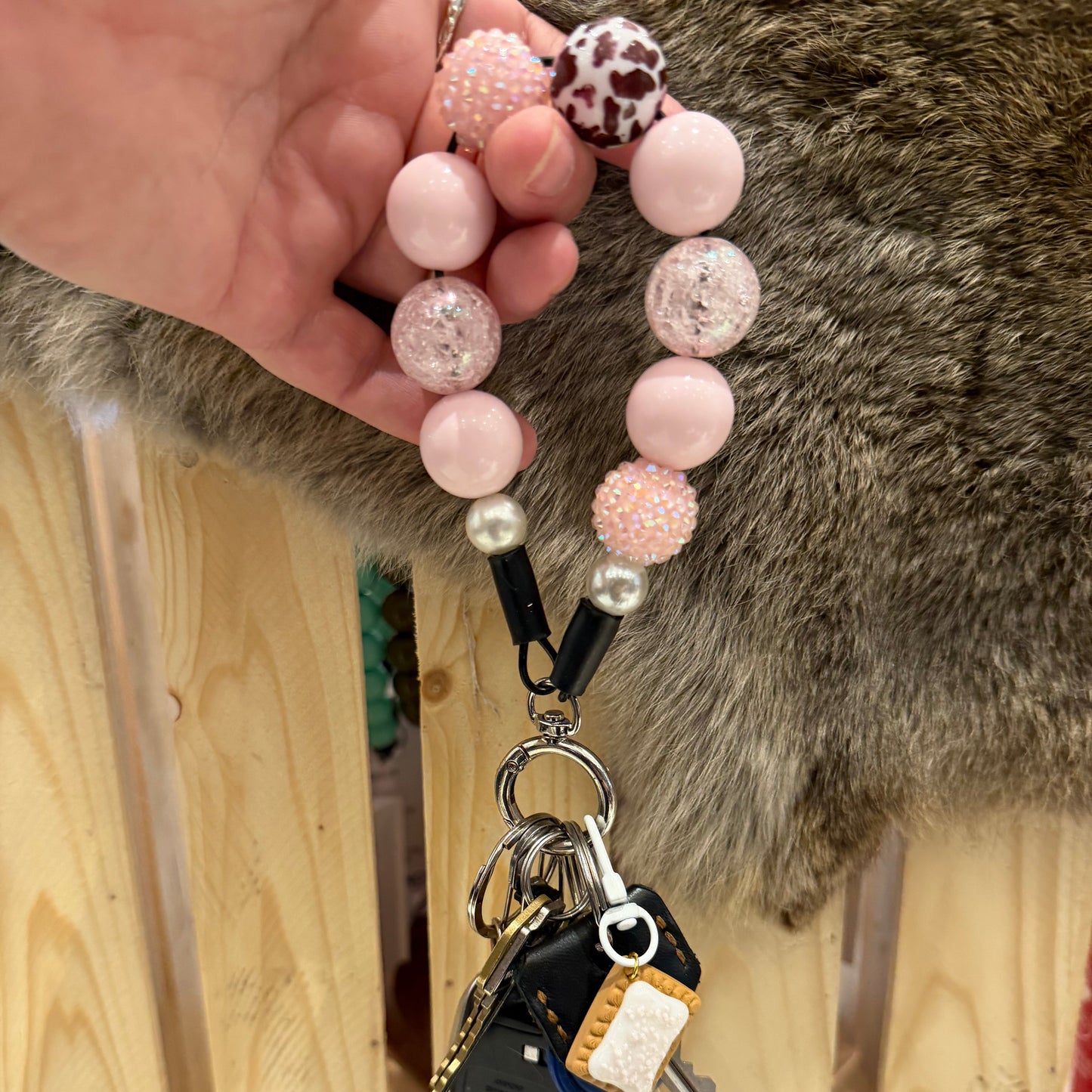 A close-up of a hand holding a pink beaded keychain with a black car key fob attached, set against a background of a wooden fence and a gray-brown furry surface.