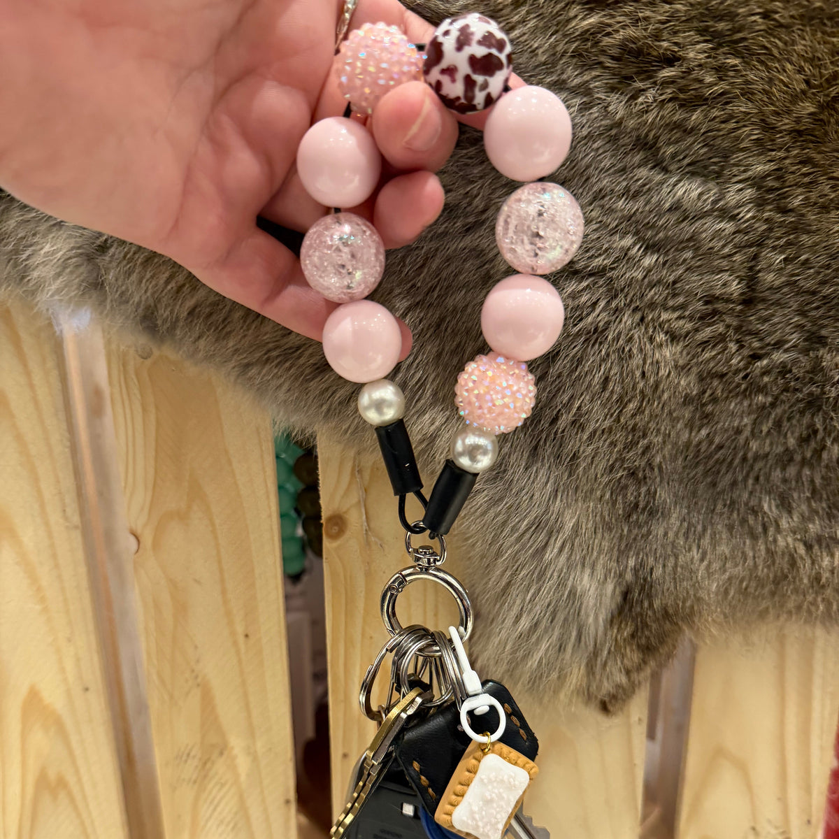 A close-up of a hand holding a pink beaded keychain with a black car key fob attached, set against a background of a wooden fence and a gray-brown furry surface.