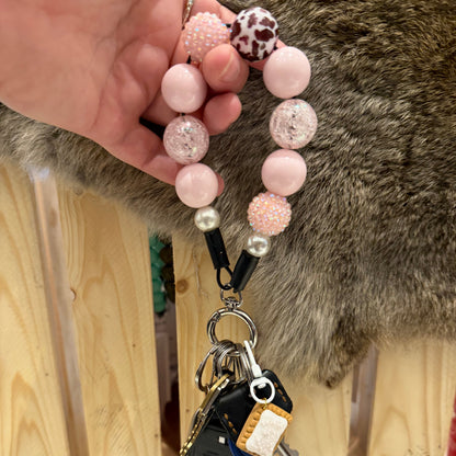 A close-up of a hand holding a pink beaded keychain with a black car key fob attached, set against a background of a wooden fence and a gray-brown furry surface.