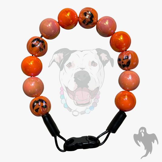 A beaded dog collar featuring glossy orange and pinkish-orange beads with black jack-o'-lantern faces, completed with a black plastic clasp. A faint graphic of a pitbull dog wearing the collar is displayed in the background.