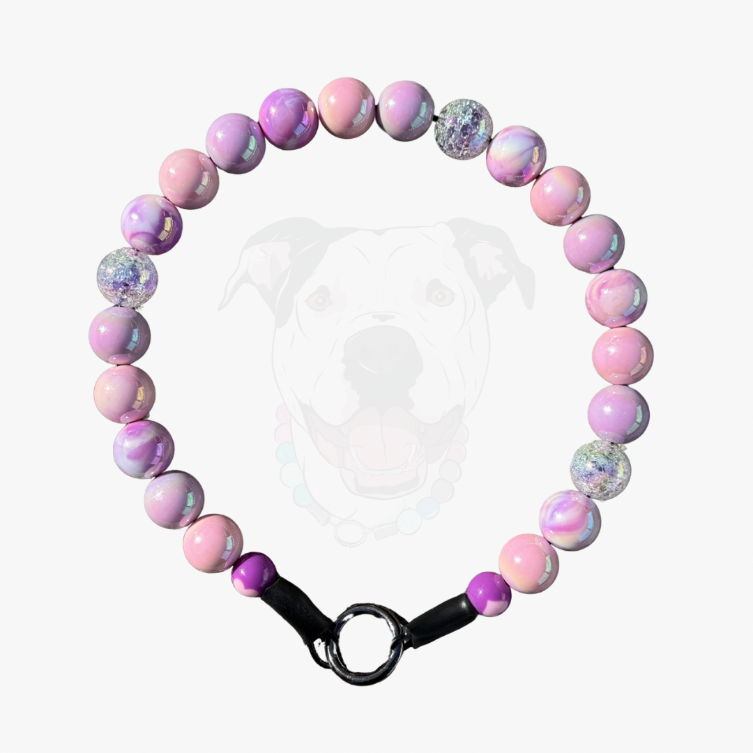 A beaded dog collar featuring pastel pink, lavender, and iridescent marbled beads, with a black metal ring clasp, displayed against a light gray background with a faint dog silhouette.