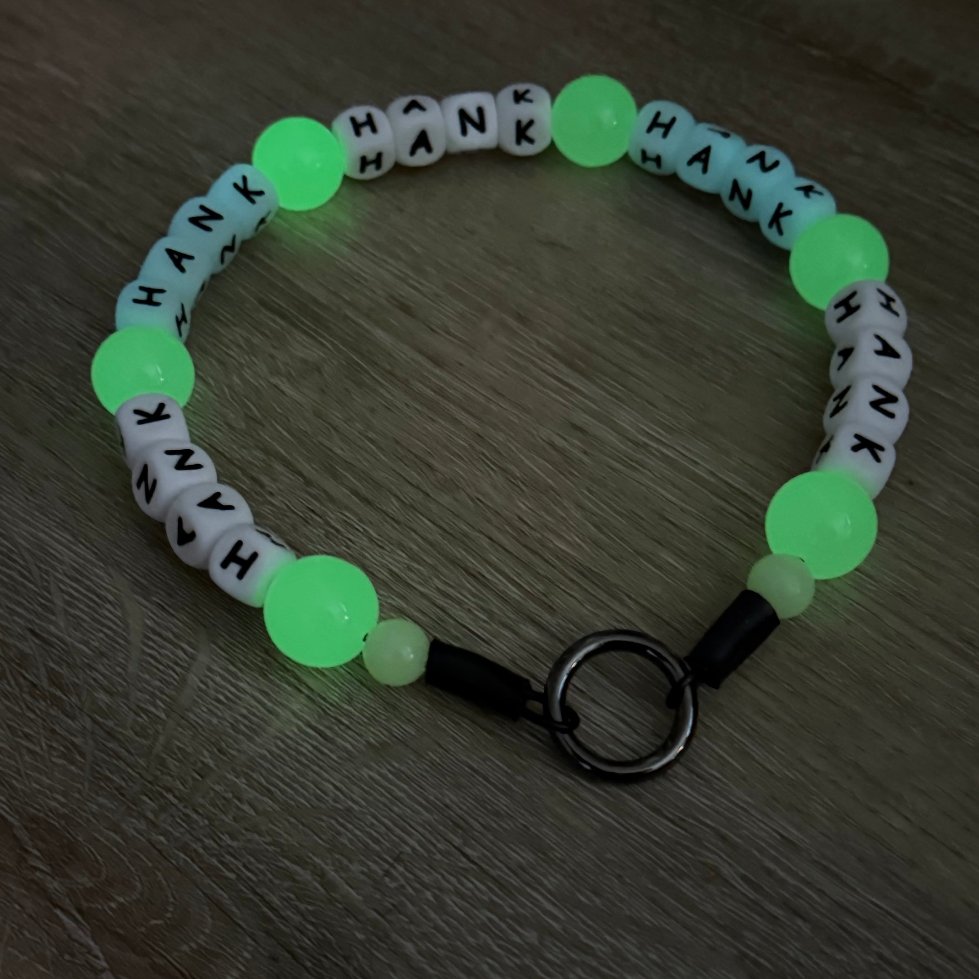Close up of the Liora Custom Glow in the Dark POPNCO Beaded Pet Collar on a wood floor. The collar features repeating 12mm silicone beads that spell out HANK and between each Hank is a 20mm glow in the dark green bead