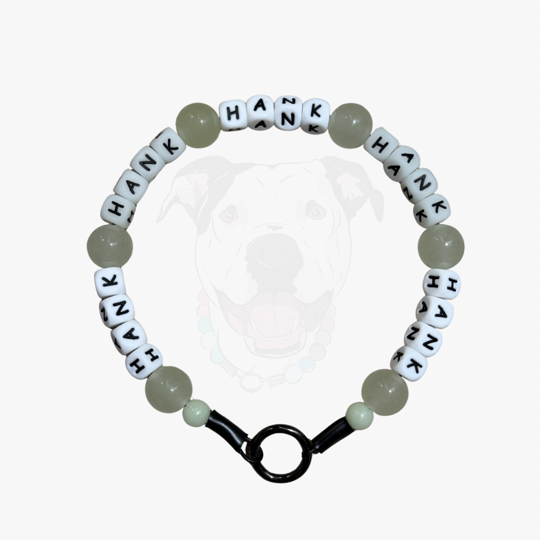A beaded dog collar featuring white letter beads spelling 'HANK' repeatedly, light green round beads, and a black clasp, shown against a light gray background with a faint dog graphic.