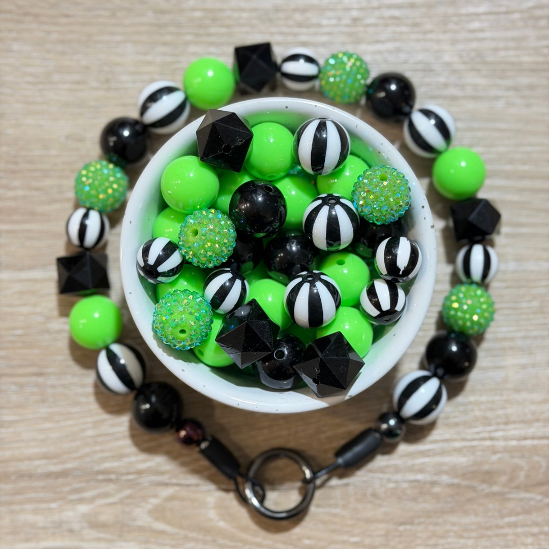 A beaded pet collar arranged in a circular shape around a white bowl filled with matching beads. The beads are primarily neon green, glossy black, and black-and-white striped, with some glittery green beads and faceted black geometric charms.