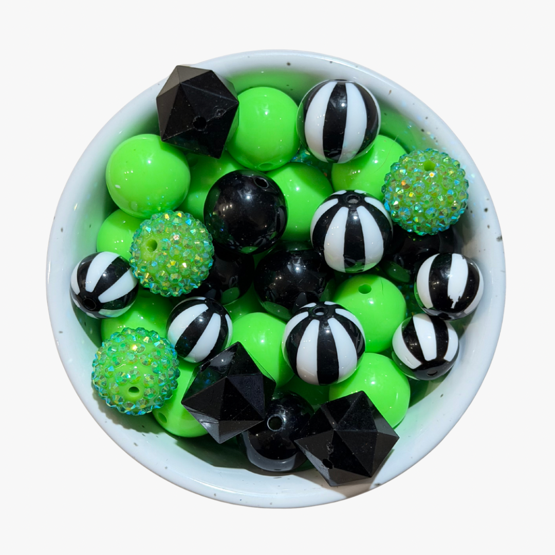 A top-down view of a white speckled bowl filled with assorted craft beads, including solid neon green, black, white, and striped black-and-white beads, some with iridescent glitter.