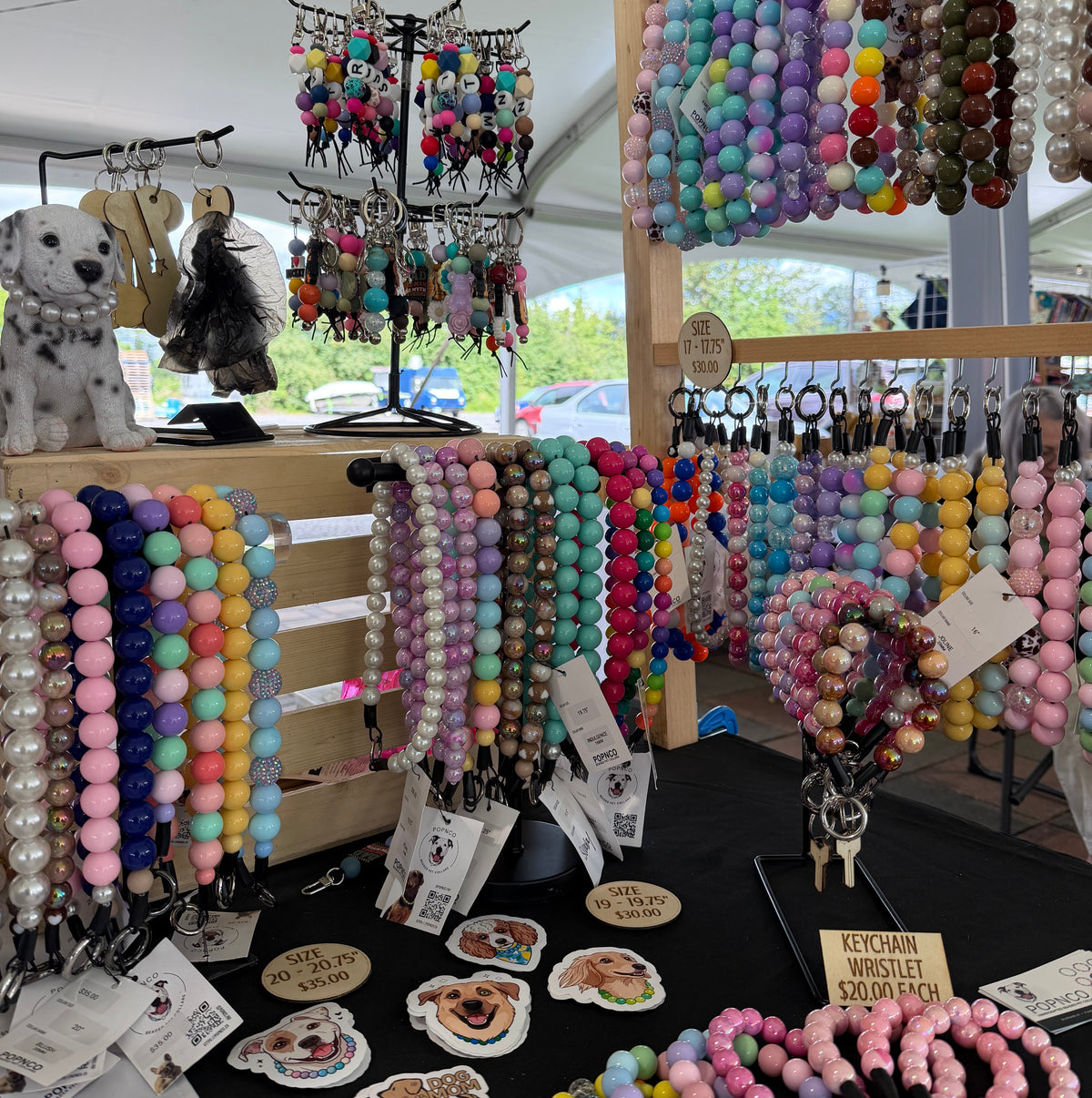 A market stall display of colorful beaded dog accessories, including keychains, bracelets, and dog-shaped stickers, under a white tent.