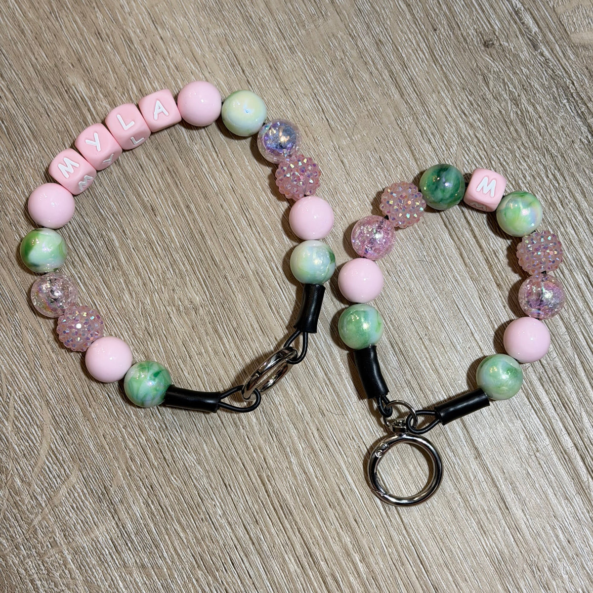 Two personalized beaded bracelets arranged on a light wood floor. One bracelet spells 'MCLA' in letter beads, and the other has 'M' and 'W' letter beads.
