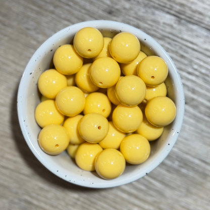 A top-down view of glossy yellow round beads with small holes, arranged in a white speckled ceramic bowl on a light gray wood-grain surface.