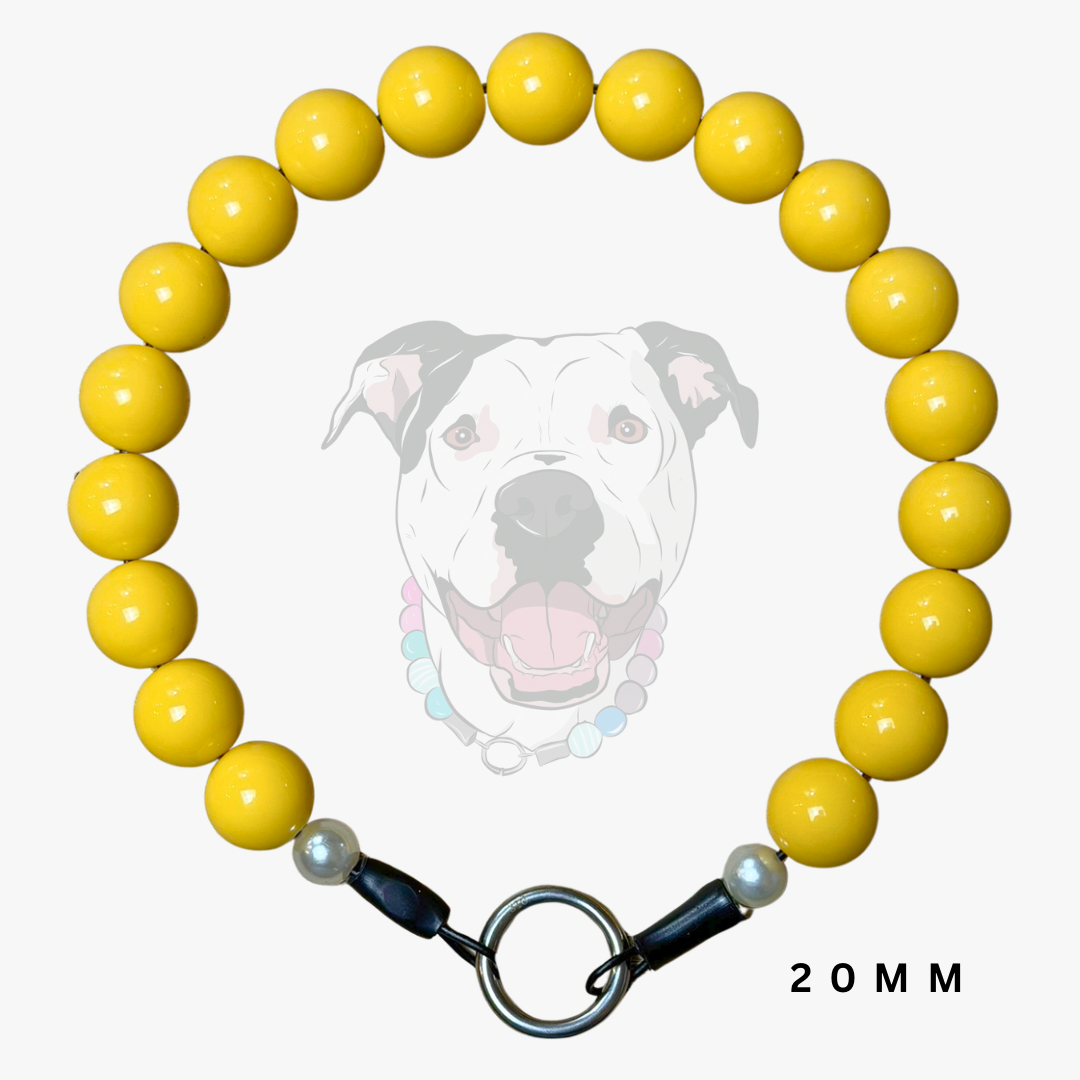A beaded dog collar made of glossy yellow round beads, featuring a silver metal ring clasp, black and white toggle ends, and a small white pearl accent. A faint graphic of a dog's face is visible in the background, and the text '20 MM' is printed in the bottom right corner.