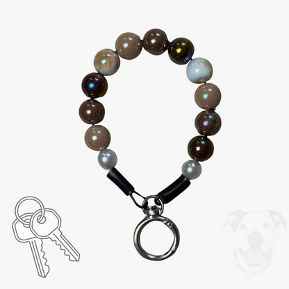 A beaded keychain with iridescent brown and white beads, black leather loops, and a silver metal split ring, shown on a light gray background with a small key graphic and a faint dog silhouette.