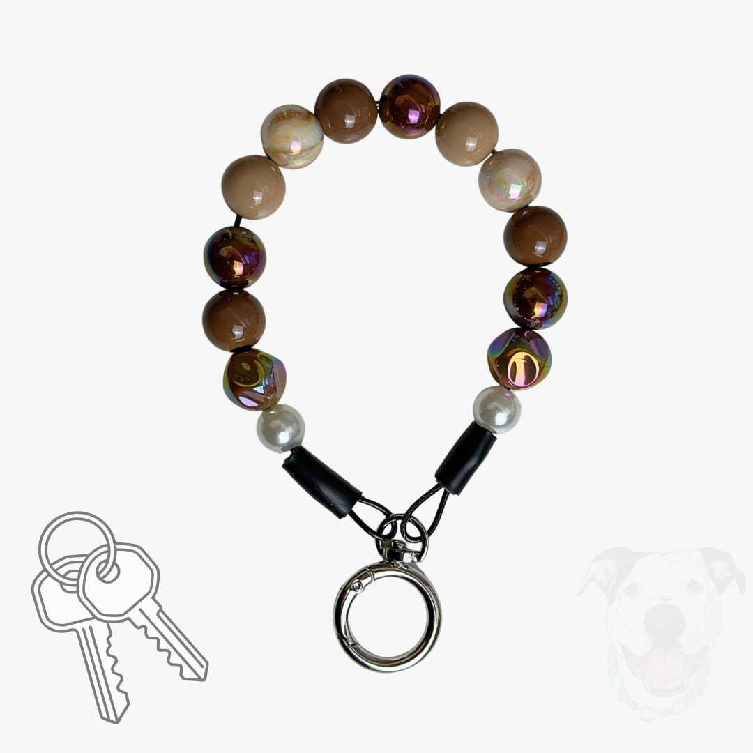 A beaded keychain with brown and iridescent beads, white pearl-like accents, and a silver split ring, shown on a light gray background with small decorative graphics of keys and a dog silhouette.
