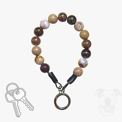 A beaded dog collar or keychain-style accessory with round marbled beads in brown, cream, and iridescent tones, black connector pieces, and a silver metal ring clasp. A small key graphic appears at lower left, and a faint dog silhouette appears at lower right, on a plain light background.